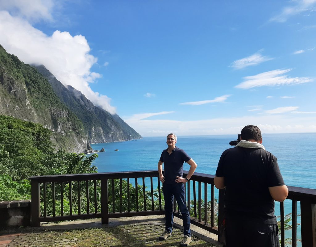 Auf einer Aussichtsplattform über dem Meer vor einer Bergkulisse steht ein Mann und wird von einem Kameramann gefilmt
