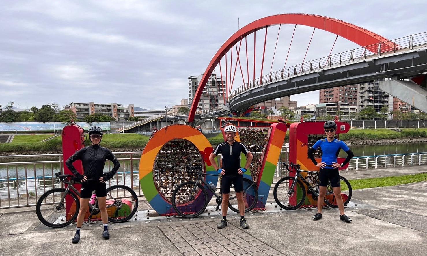 Drei Radfahrer posieren stolz vor einer Brücke am Flussufer.