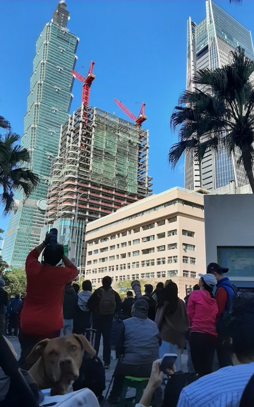 Menschen stehen auf dem Bürgersteig und schauen hoch zum Taipei 101. Einige fotografieren mit langen Objektiven.